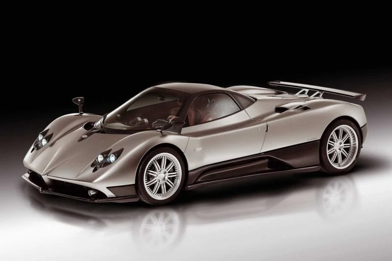 A silver sports car with aerodynamic design, large rear wing, and low profile, reminiscent of Pagani to showcase four hypercar icons at the upcoming 9th Historic Minardi Day in Imola, photographed on a reflective surface against a dark gradient background.