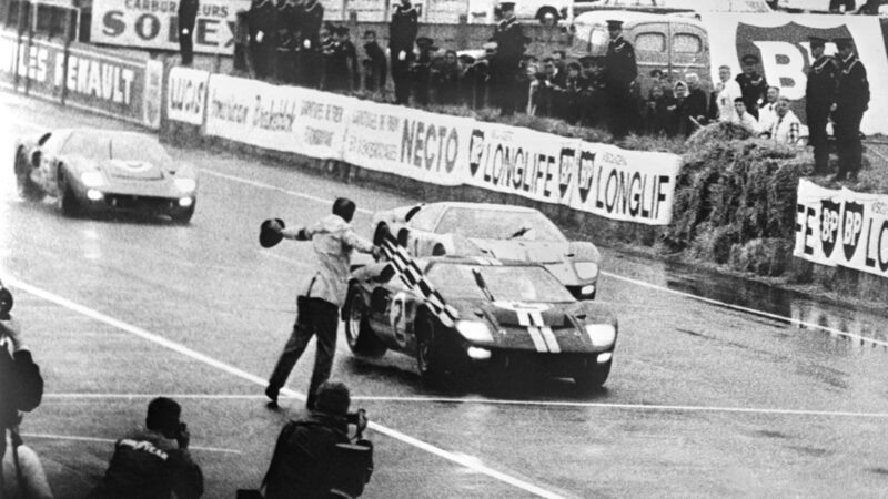 Two race cars cross the finish line on a rainy track at Le Mans 2027 as a man waves a flag, with spectators watching from behind barriers—capturing the excitement of Ford vs Ferrari.