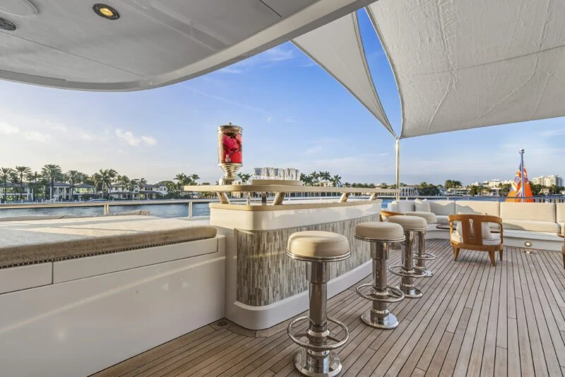 Experience the elegance of a luxury yacht deck aboard Delta Marine Noverra, featuring a shaded bar area with four bar stools, a large glass beverage dispenser, and breathtaking views of palm trees and tranquil water.