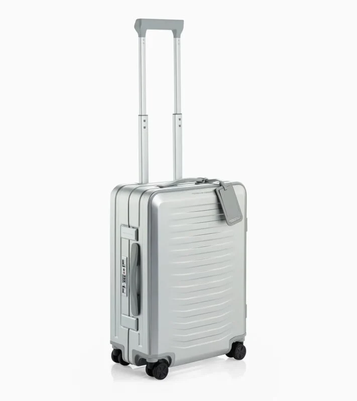 A silver Porsche Design hard-shell carry-on suitcase with four wheels, an extended telescopic handle, and a travel tag attached to the top—an ideal pick for your 2025 Holiday Gift Guide.