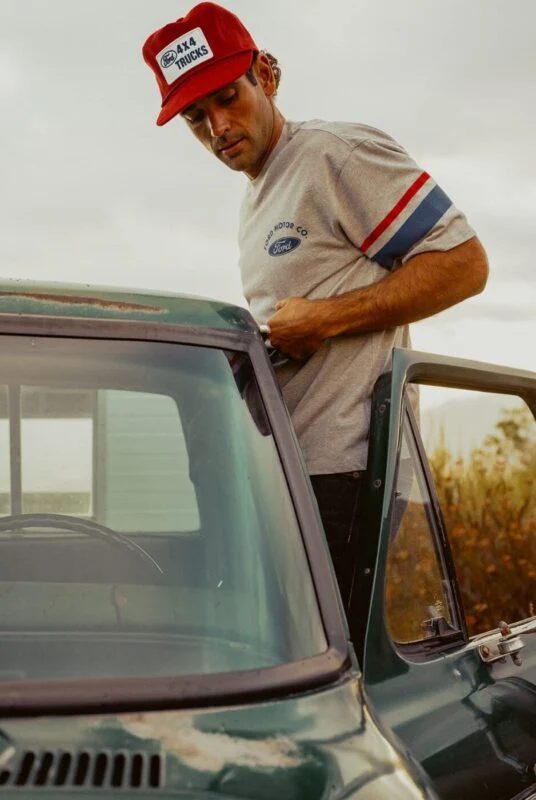 A man wearing a red cap and gray t-shirt stands beside an open door of a green vintage truck from the Ford x Brixton Limited Edition Collection, looking down with an overcast sky in the background.