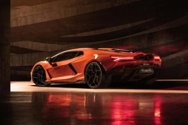 A sleek orange Lamborghini Revuelto with an angular design and glowing taillights is parked on a reflective floor against a dark, textured background, showcasing its cutting-edge specs.