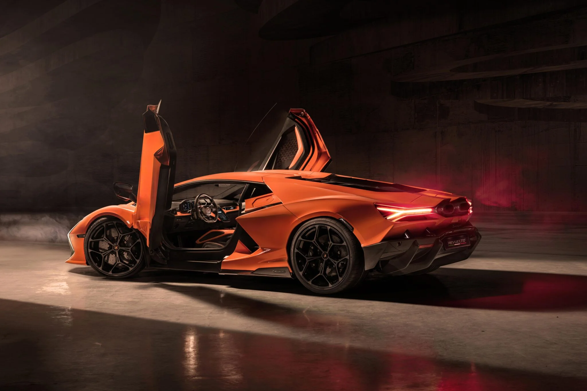 In low lighting, the Lamborghini Revuelto sports car stands with its scissor doors open, showcasing its sleek design and alloy wheels while the illuminated tail lights accentuate its elegance.