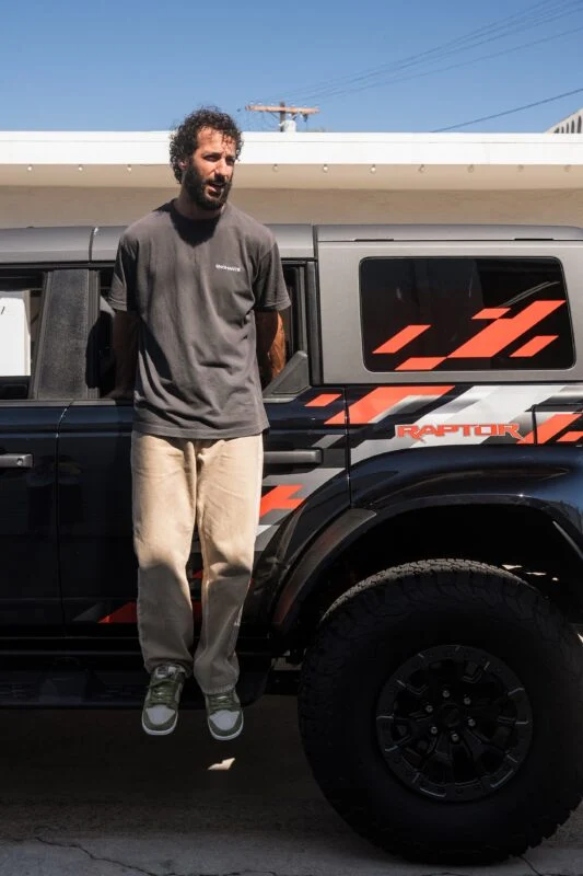 A man in a gray t-shirt and beige pants sits on the side step of a black Ford Performance Raptor with orange decals on a sunny day.