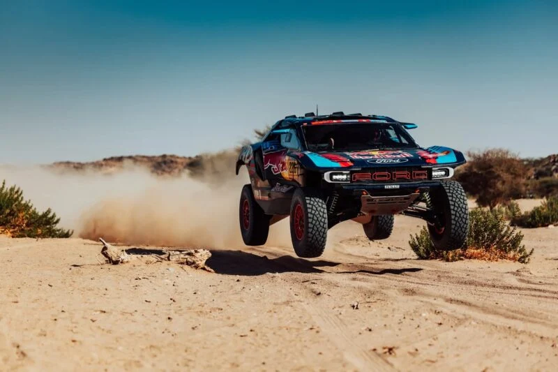 A Ford rally truck with Red Bull branding is airborne on a dusty desert track, kicking up a large cloud of dust behind it.