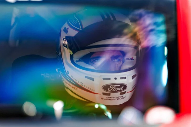 Close-up of a race car driver wearing a white Ford helmet, seen through a car window with colorful reflections—a nod to the spirit of Two Fords EVs Two Records: Lightning SuperTruck and SuperVan conquer the Green Hell.