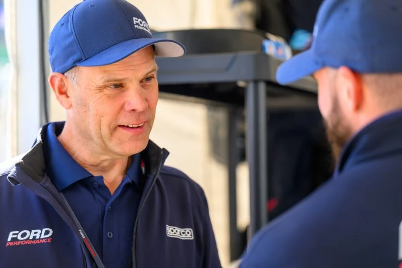 Two men wearing blue Ford Performance jackets and hats discuss indoors. As they talk, the moment captures excitement around the Ford Performance Launches Final Production Run of Legendary Ford GT Mk IV. One man faces the camera; the other has his back turned.