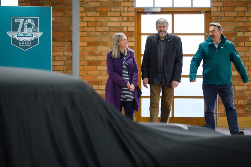 Three people stand and smile near a covered car, possibly a rare DB5, inside a building with brick walls and large windows; an Aston Martin Works 70th anniversary sign is visible.