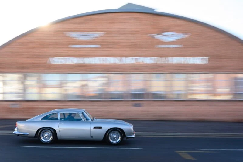 A rare DB5 silver classic car drives past a red brick building with 