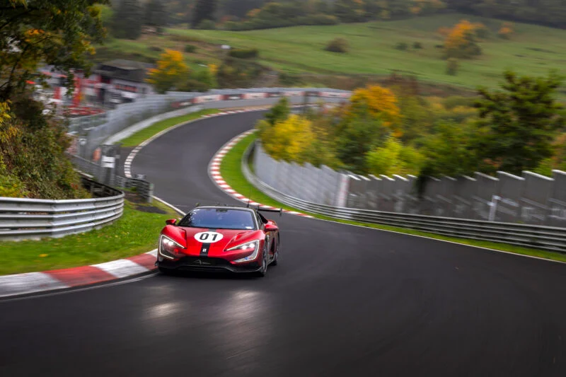 A red electric hypercar marked with number 07 drives around a sharp corner on a racetrack surrounded by greenery and trees, reminiscent of a Nürburgring record attempt.