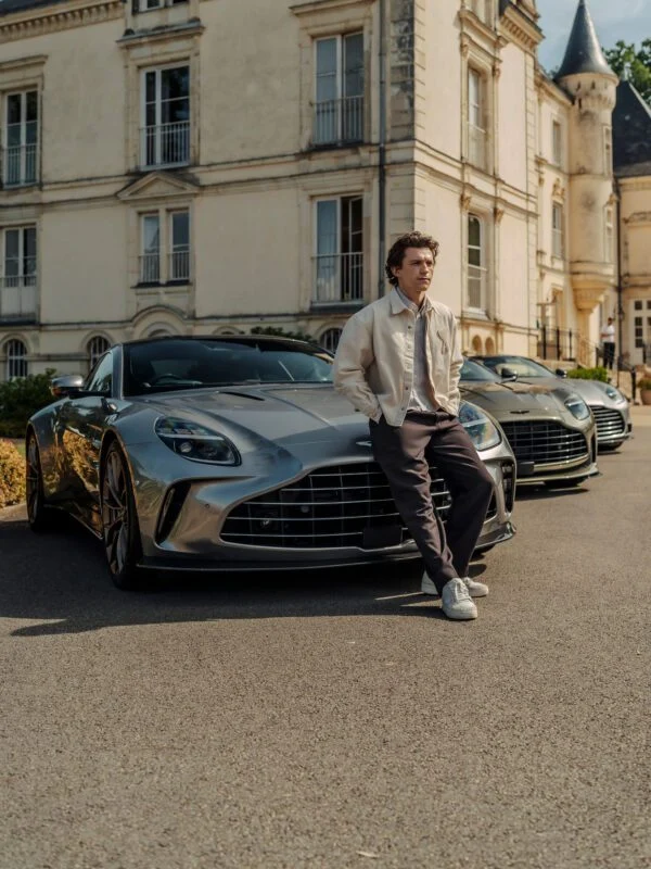 A man in a light jacket and dark pants leans against the front of a silver Aston Martin, with two similar cars and a large historic building in the background.