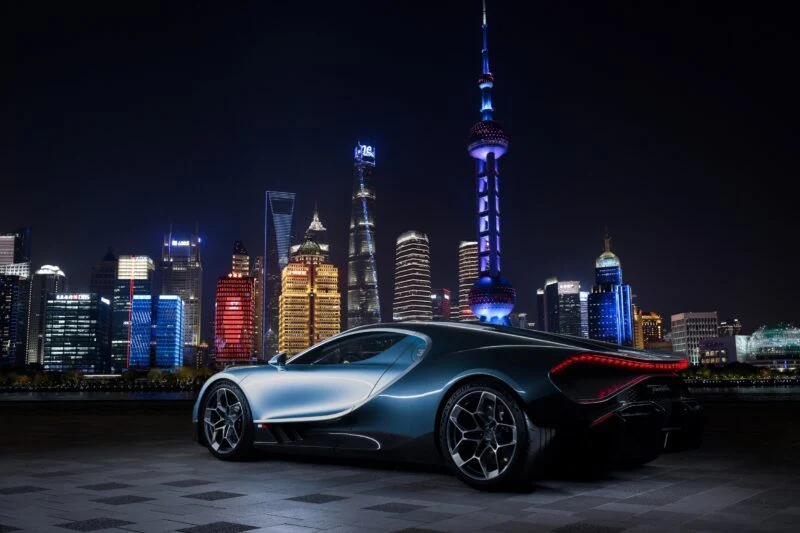 A sleek Bugatti sports car parked on a tiled surface at night, with a brightly lit modern Shanghai showroom and city skyline in the background.