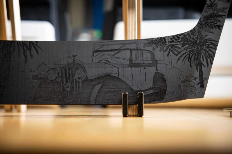 Close-up of a metal sheet engraved with a vintage car scene, featuring two people in the car and palm trees in the background, propped upright—a tribute as Rolls-Royce honors 100 years of Phantom with groundbreaking private collection art.