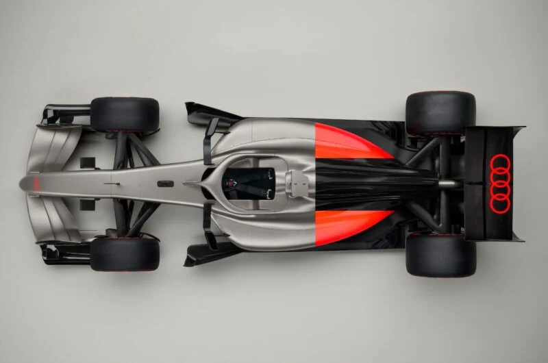 Top view of a sleek, silver Formula 1 Audi R26 concept car with black and orange accents and an Audi logo on the rear wing, photographed on a plain gray background.