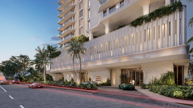 Street-level view of a modern high-rise, part of the Aston Martin Expands Real Estate Empire With Daytona Beach Shores Residences, featuring balconies, lush greenery, palm trees, and a car parked near the entrance at sunset.